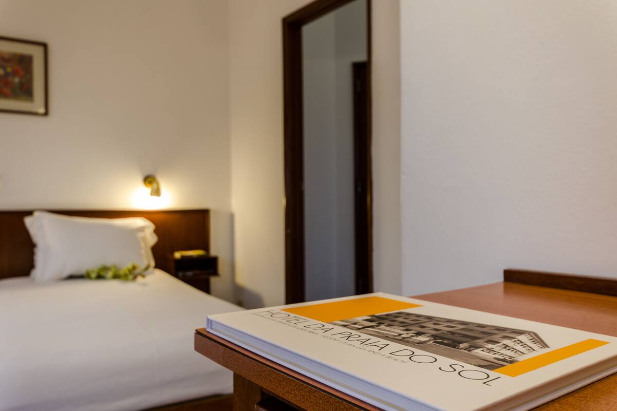 Une chambre d’hôtel avec un lit bien fait, une table de chevet sur laquelle repose un livre et une petite table de nuit avec une lampe.