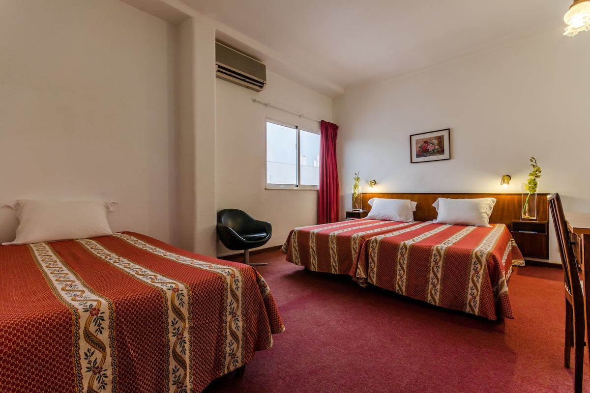 Une chambre d’hôtel avec deux lits, un bureau, une chaise, une fenêtre avec des rideaux, un climatiseur et un tableau encadré au mur.