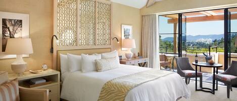 A hotel room with a large bed, two bedside tables with lamps, a chair, a small table with a bottle and glasses, and a balcony with a view of the mountains.
