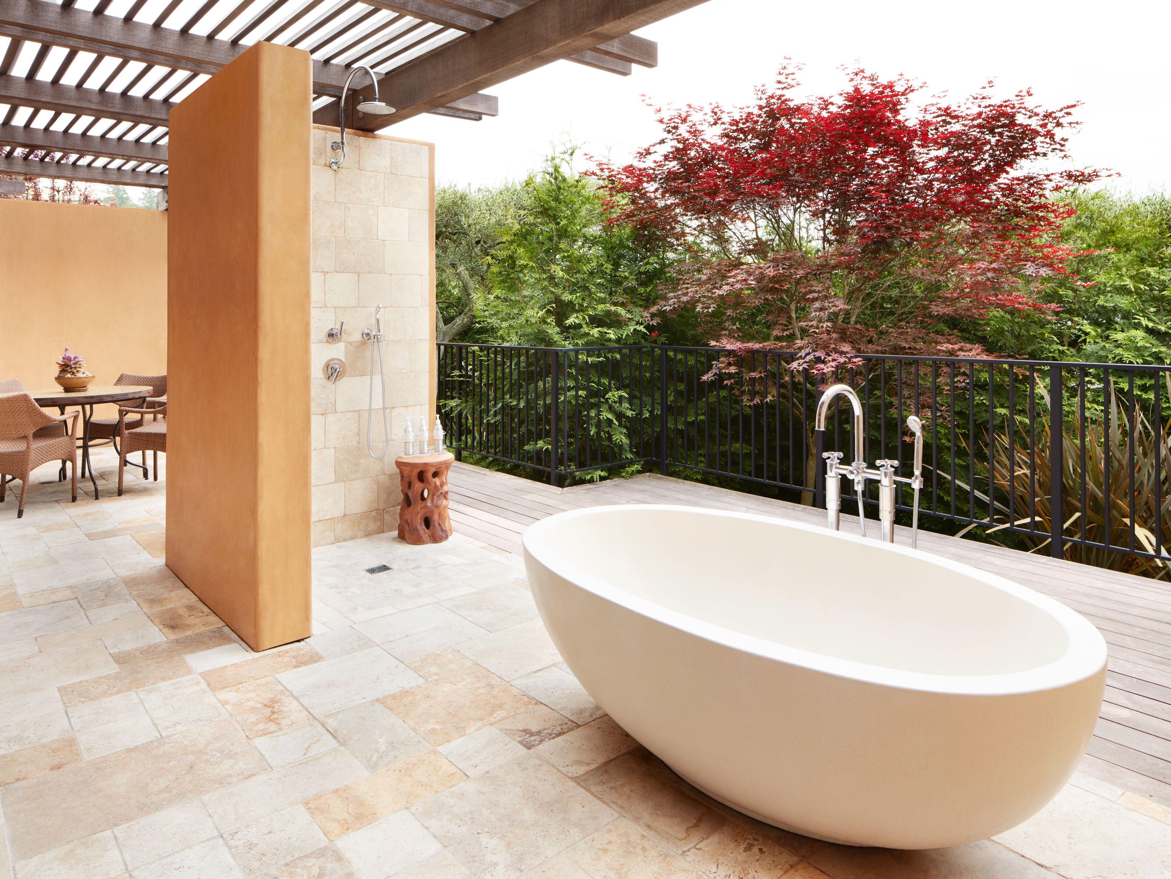 A modern outdoor bathroom with a freestanding bathtub, a seating area with chairs, and a wooden table.