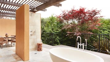 A modern outdoor bathroom with a freestanding bathtub, a seating area with chairs, and a wooden table.