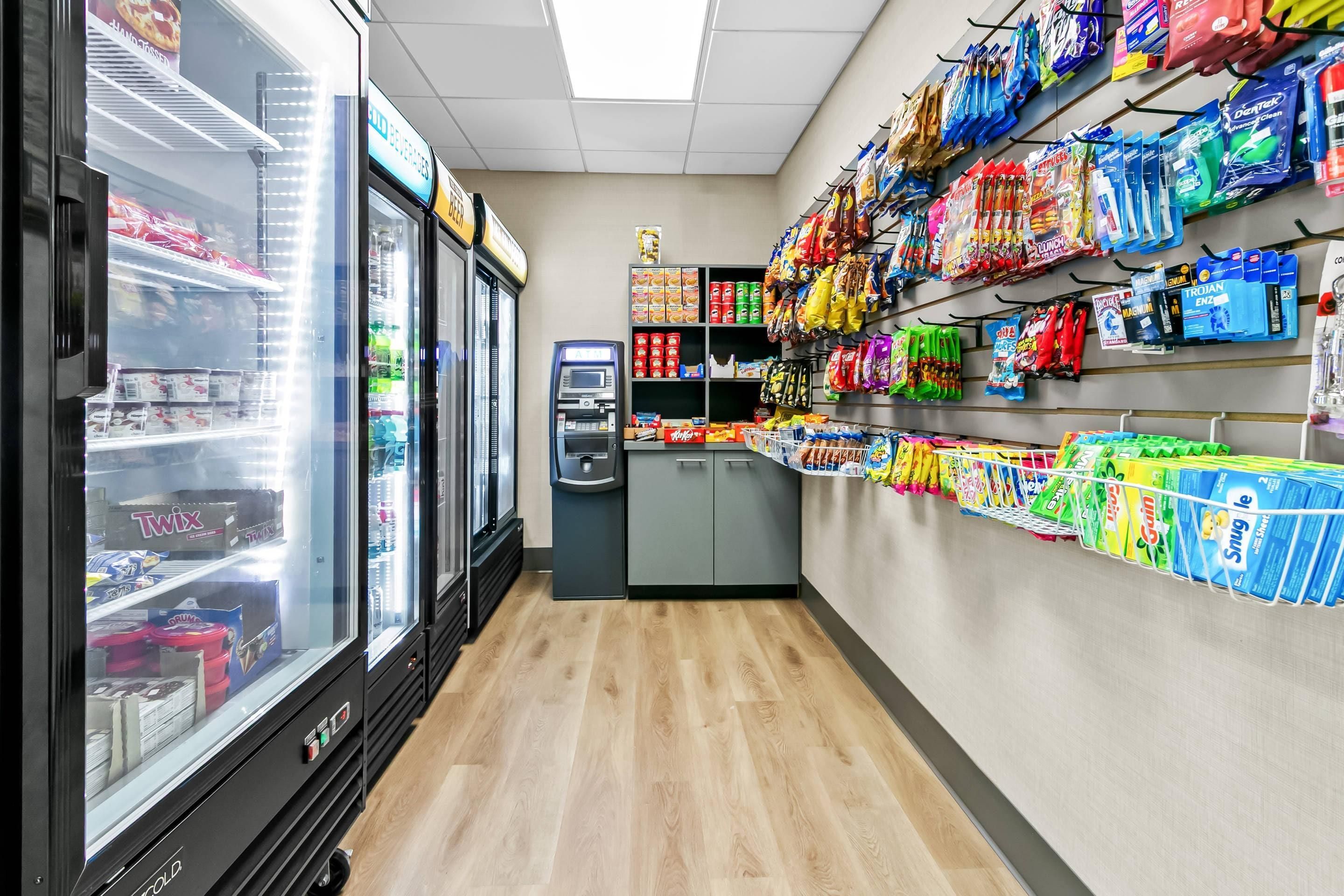 A convenience store with refrigerated freezers on the left, a self-checkout machine, and shelves stocked with snacks and household items.