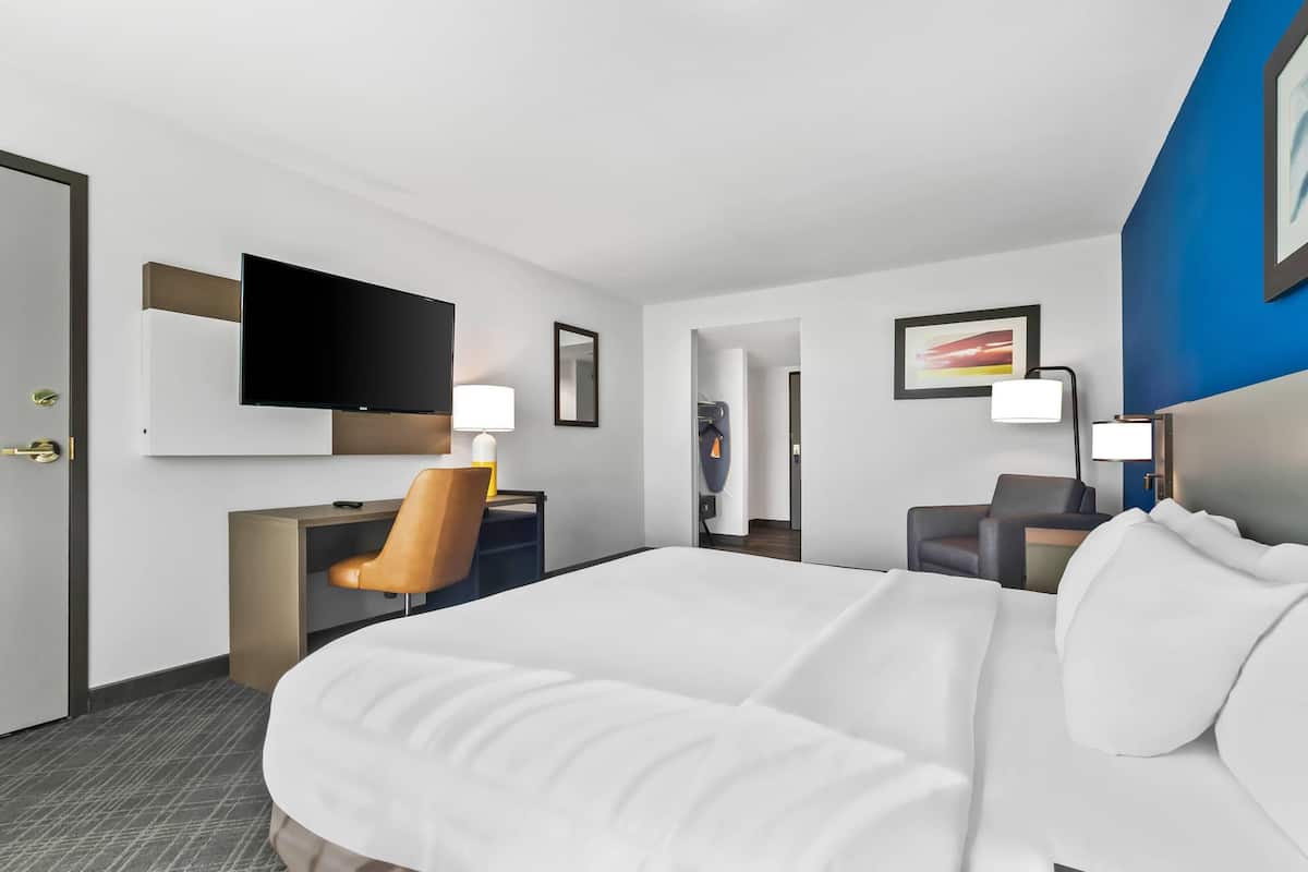 A hotel room with a large bed, a desk with a chair, a flat-screen TV mounted on the wall, and a bathroom visible through an open door.