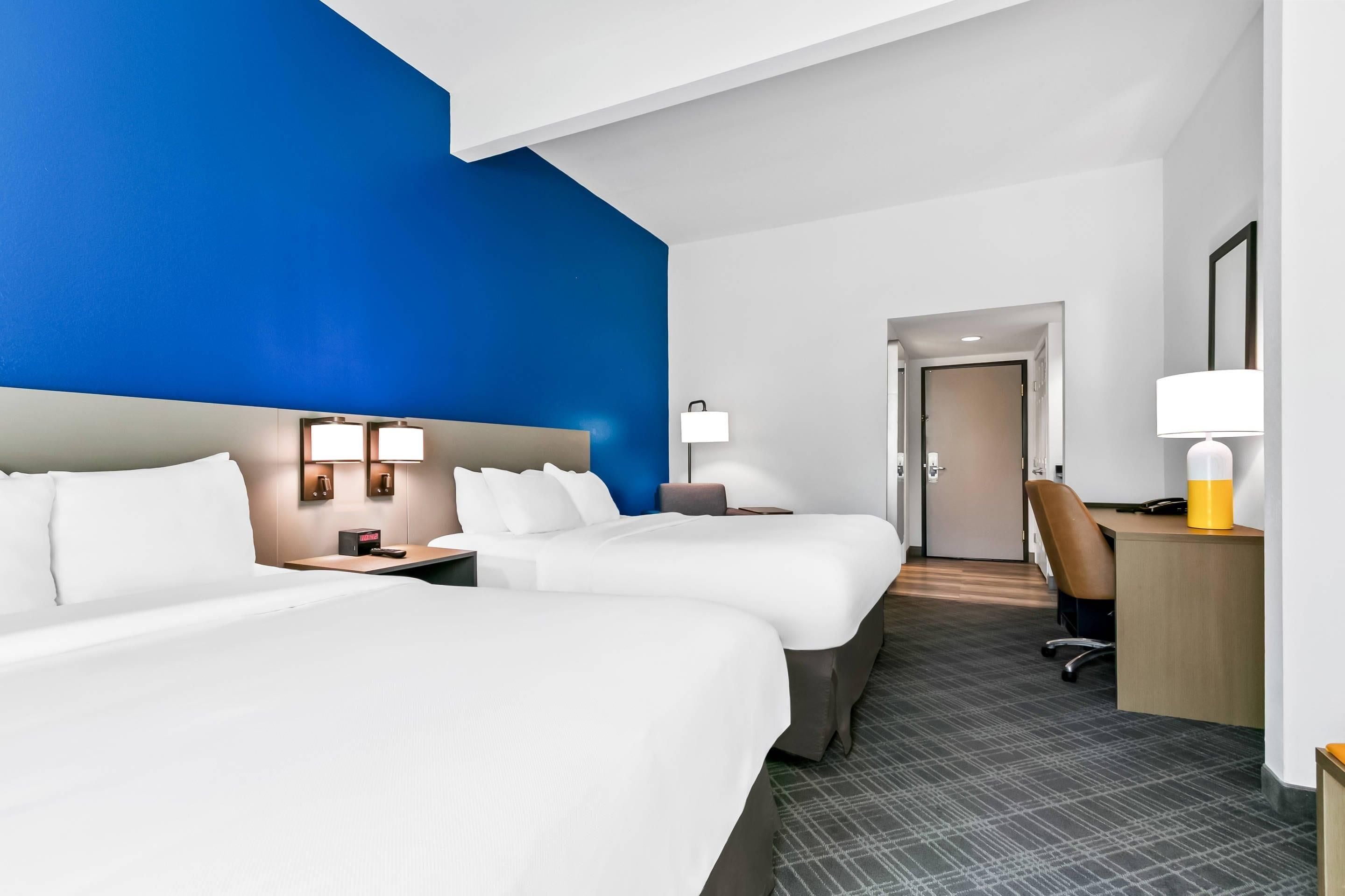 A hotel room with two beds, a desk, a chair, and a lamp.