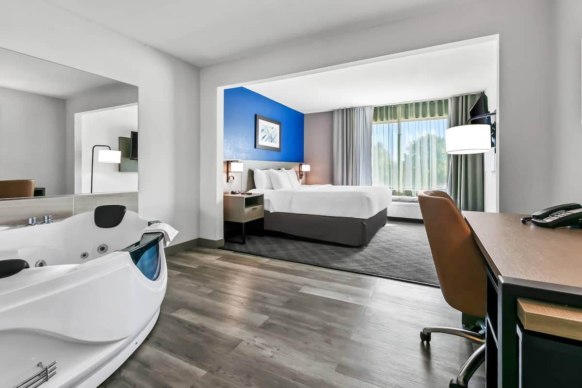 A hotel room with a large bathtub, a bed with a headboard, a desk with a chair, and a window with curtains.