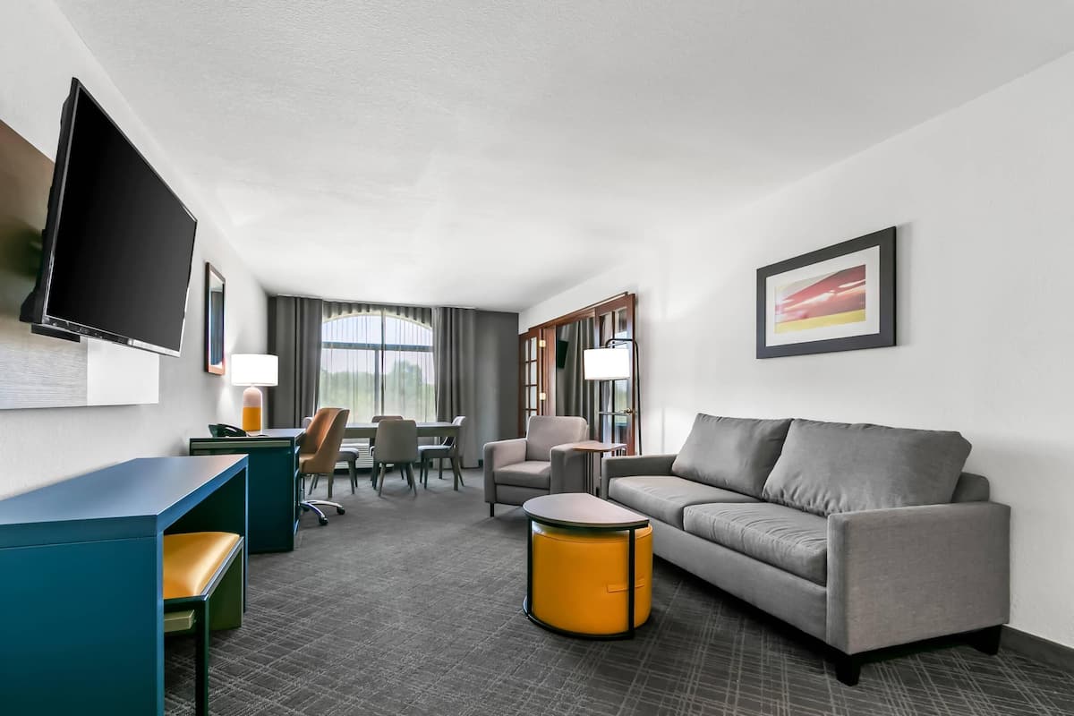 A hotel room with a grey sofa, a blue desk, a yellow ottoman, a TV, a framed picture, and a dining area with chairs and a table.
