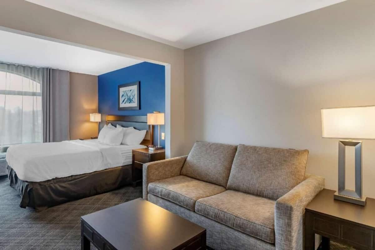 A hotel room with a bed, a sofa, a nightstand, a lamp, and a painting on the wall.