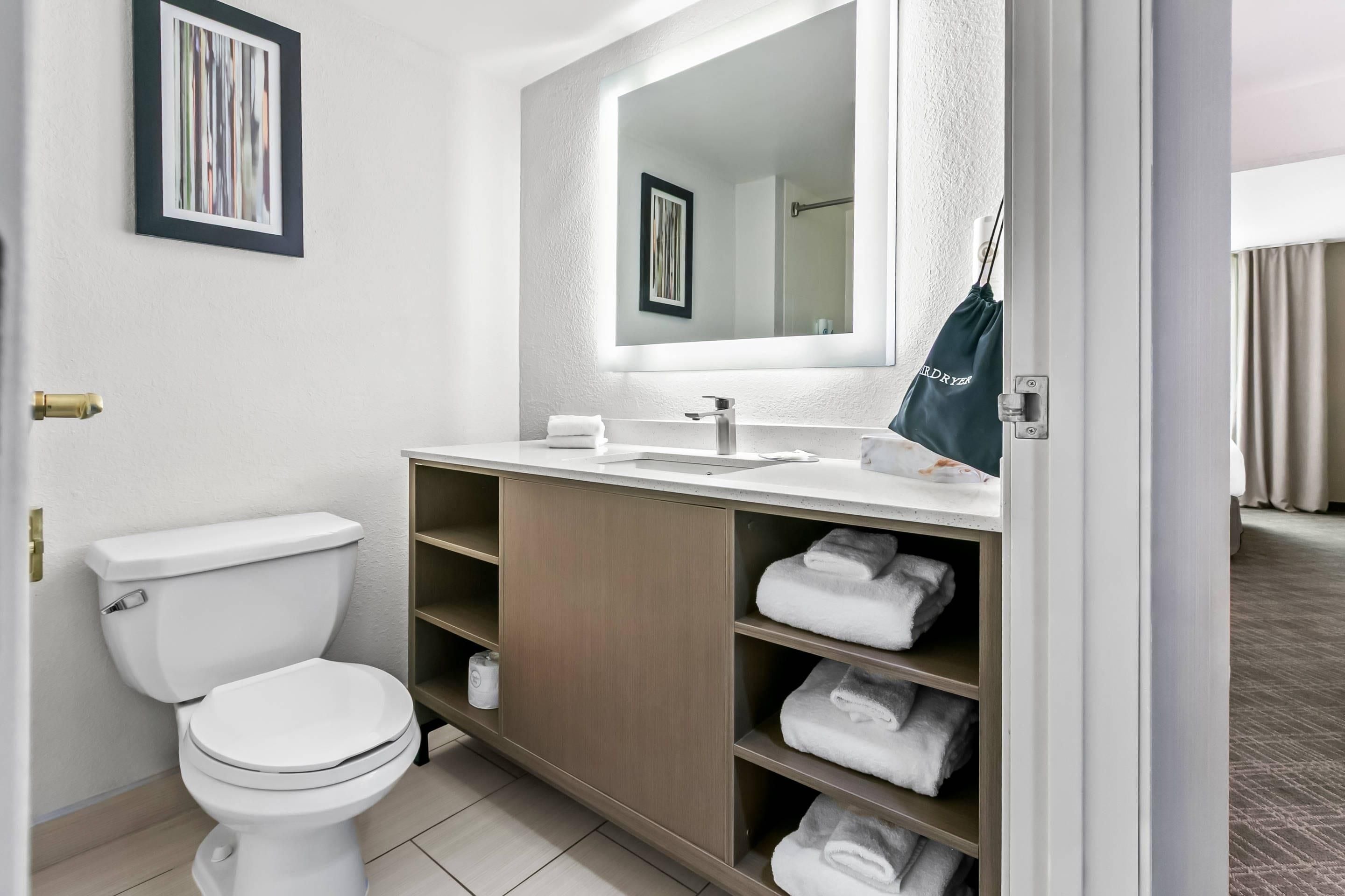 Une salle de bain moderne avec des toilettes blanches, un meuble-lavabo en bois doté d’un miroir et des étagères où sont pliées des serviettes.