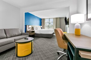 Habitación de hotel con sofá gris, una mesita de noche amarilla, cama con ropa blanca, escritorio con lámpara y vista a la ciudad por la ventana.