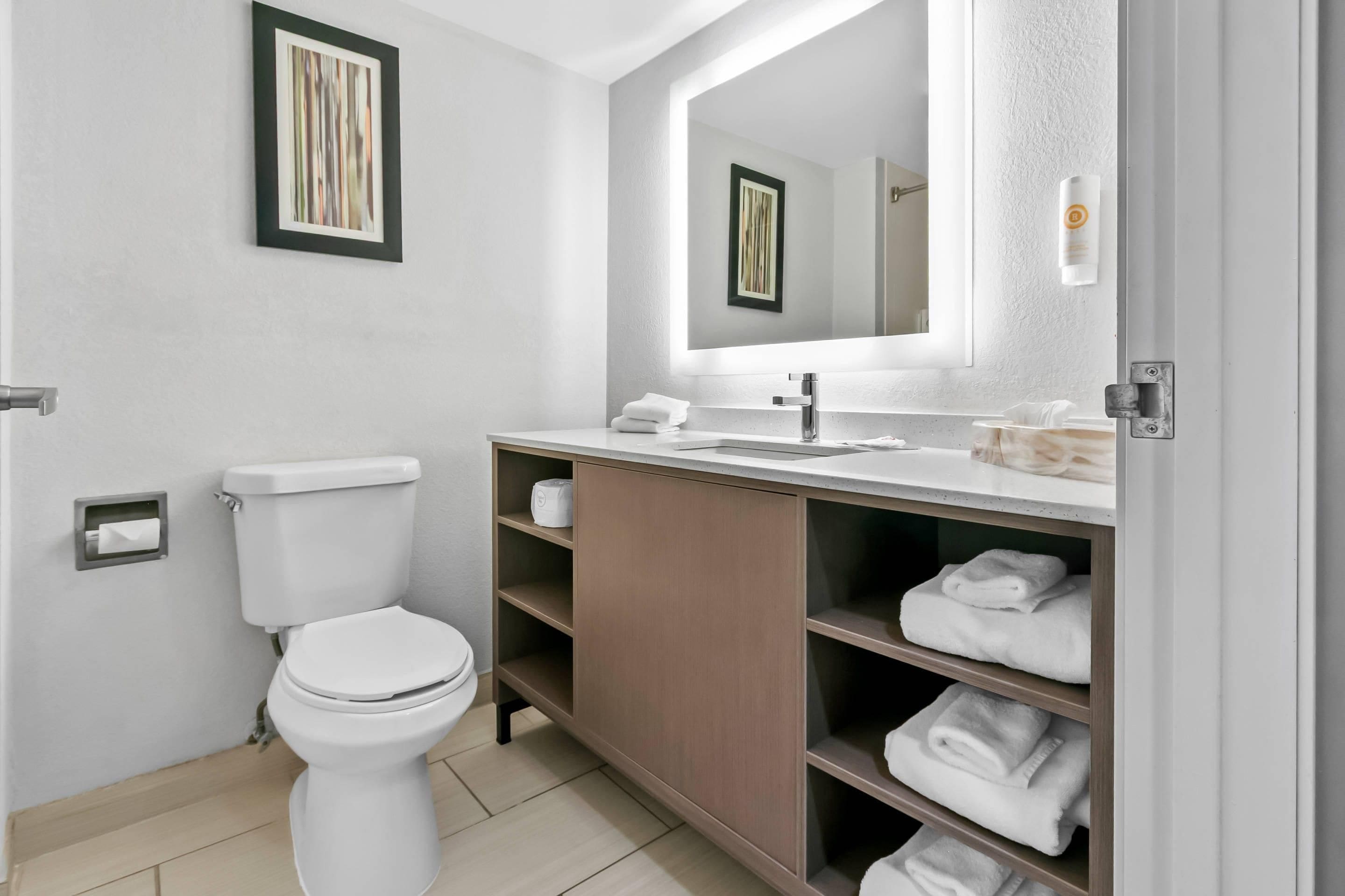 A modern bathroom with a toilet, sink, and mirror. There are shelves with towels and a framed picture on the wall.