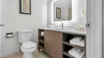 Une salle de bain moderne avec des toilettes, un lavabo et un miroir. On y trouve des étagères avec des serviettes et un tableau encadré accroché au mur.
