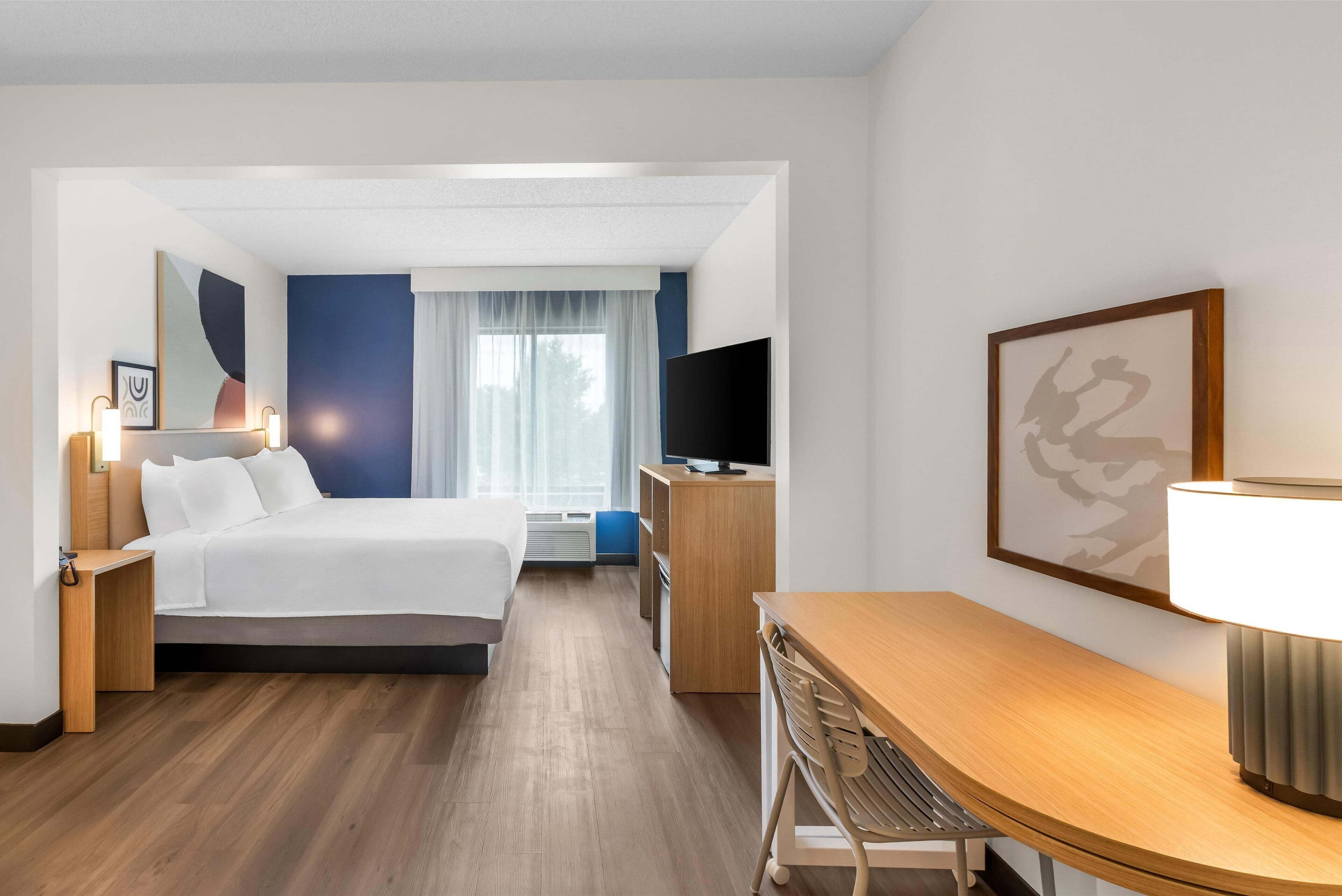 Habitació d'hotel amb llit, taula de nit, televisió, escriptori, cadira i obra d'art.