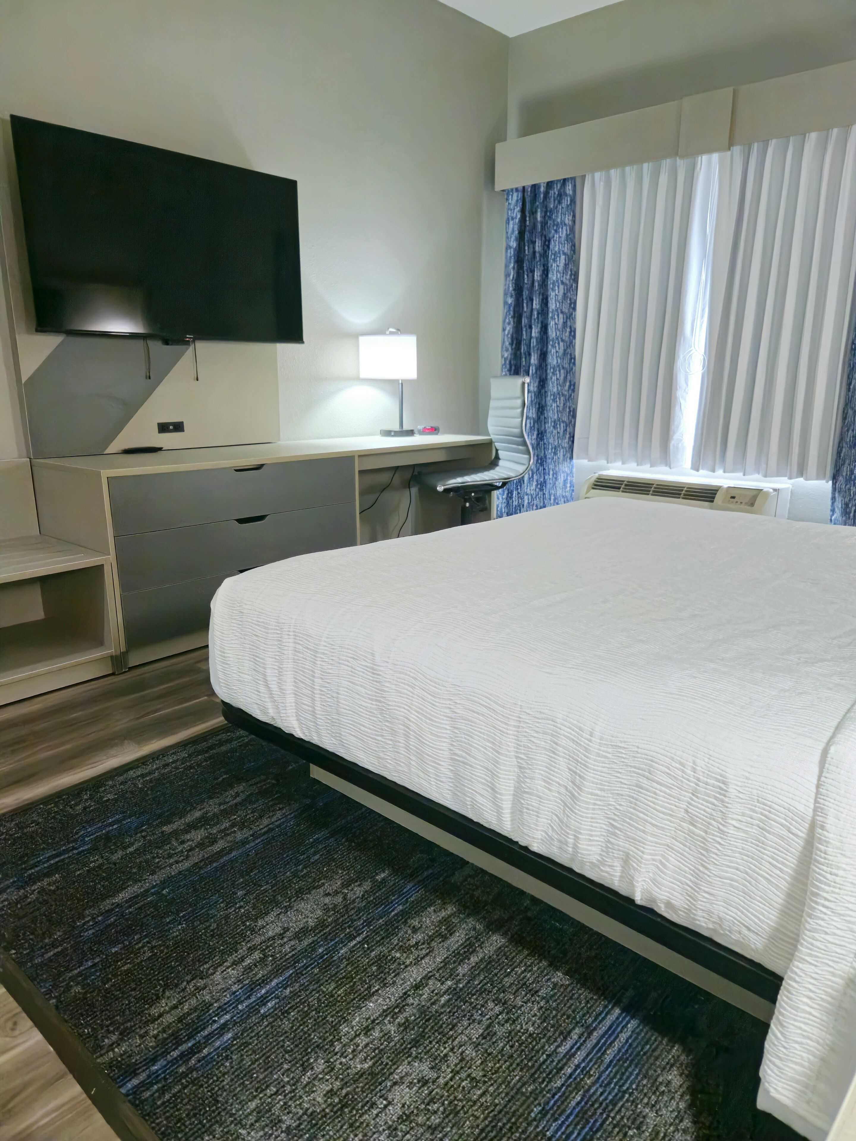 Una habitación de hotel con una cama, un escritorio, un televisor de pantalla plana, una lámpara y una ventana con cortinas.