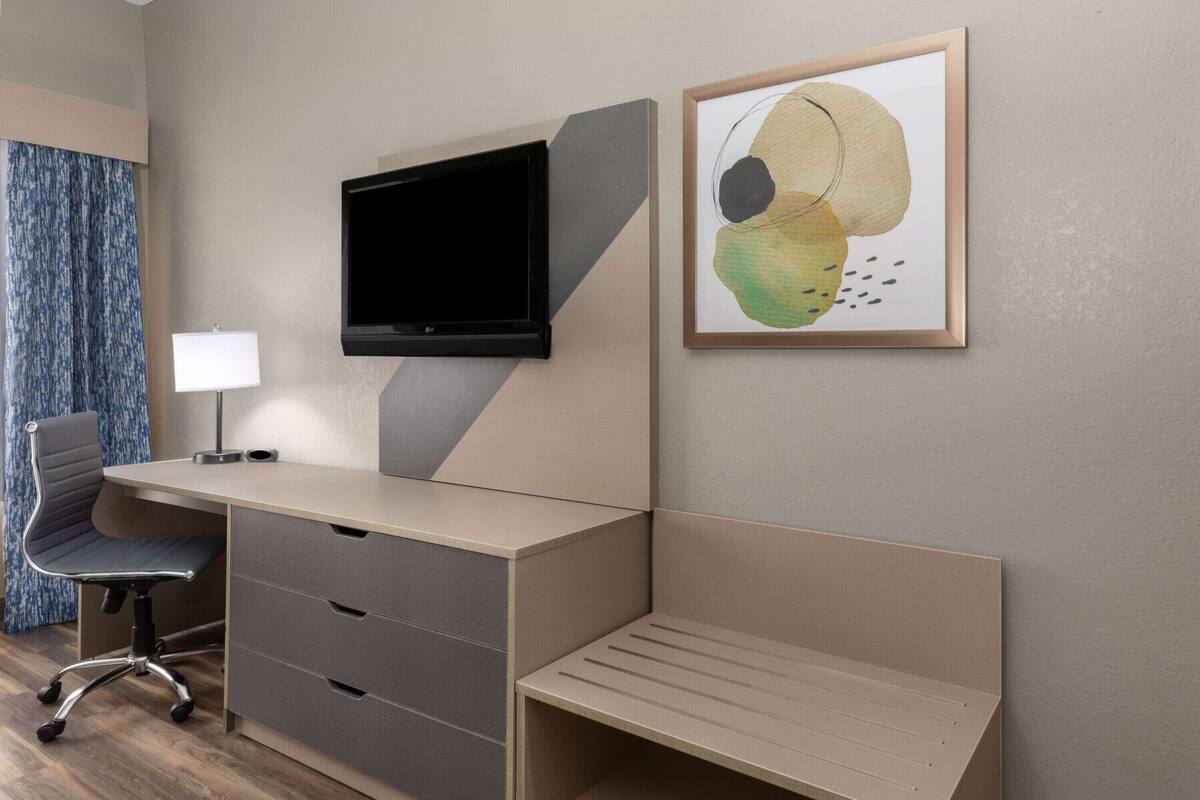A modern hotel room with a desk, chair, TV, and artwork.