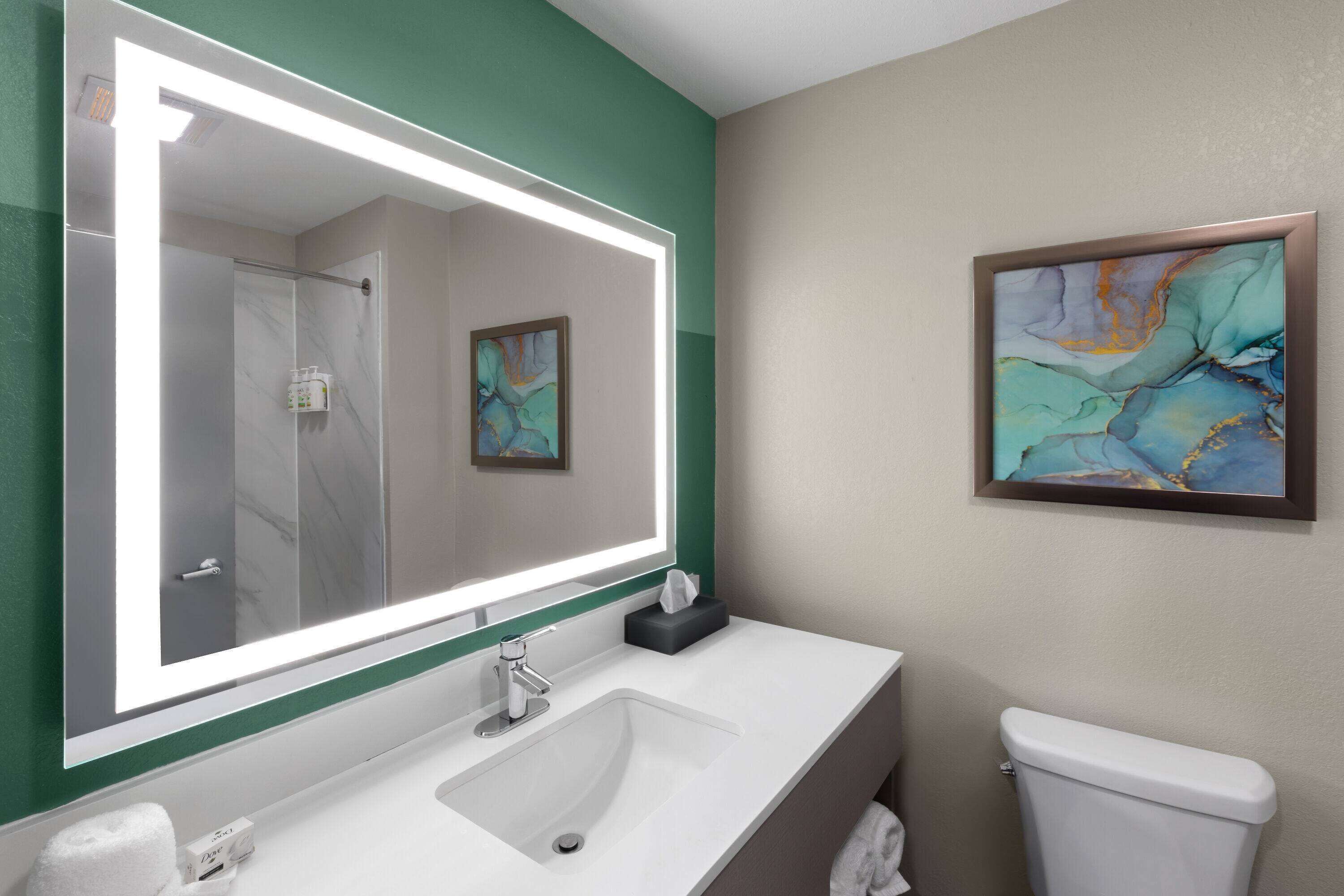 Une salle de bain avec un grand miroir, un lavabo, des toilettes et un tableau abstrait encadré accroché au mur.