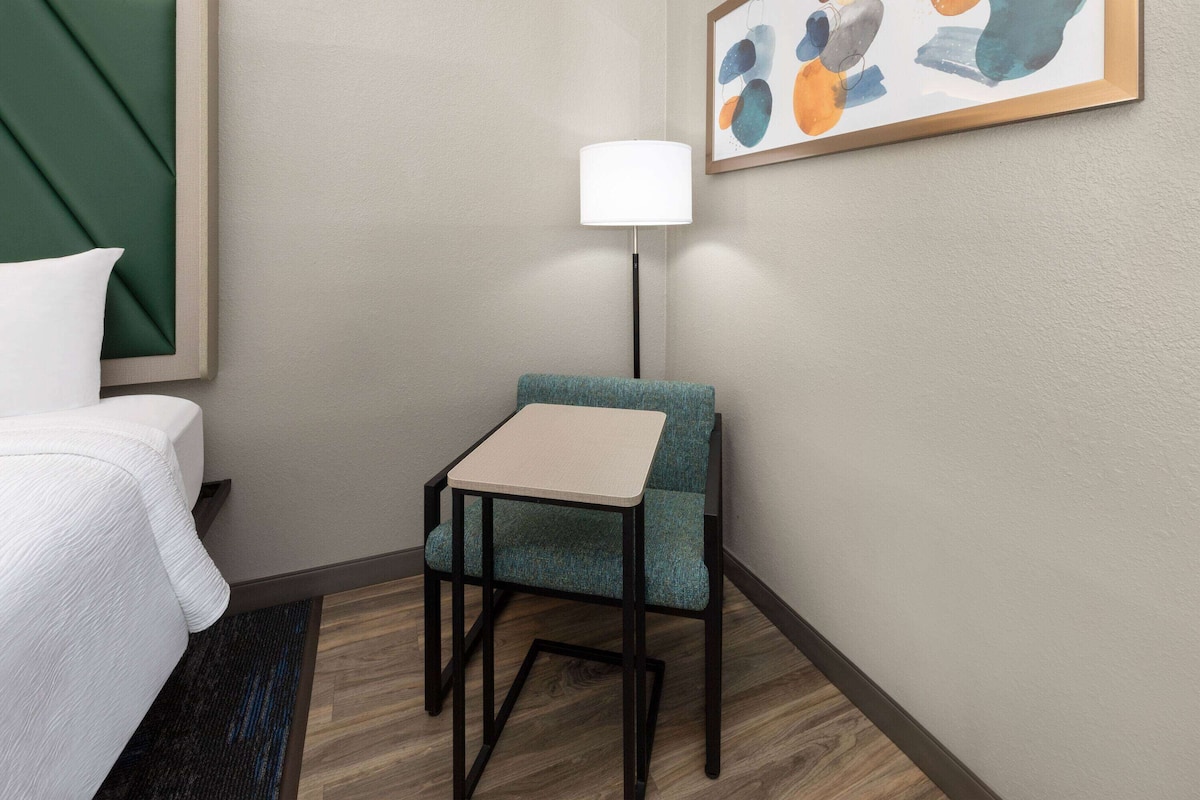 A hotel room with a bed, a small table, a chair, a floor lamp, and a painting on the wall.