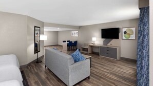 A hotel room with a bed, a grey sofa with blue cushions, a desk with a chair, a flat-screen TV, and a mirror.
