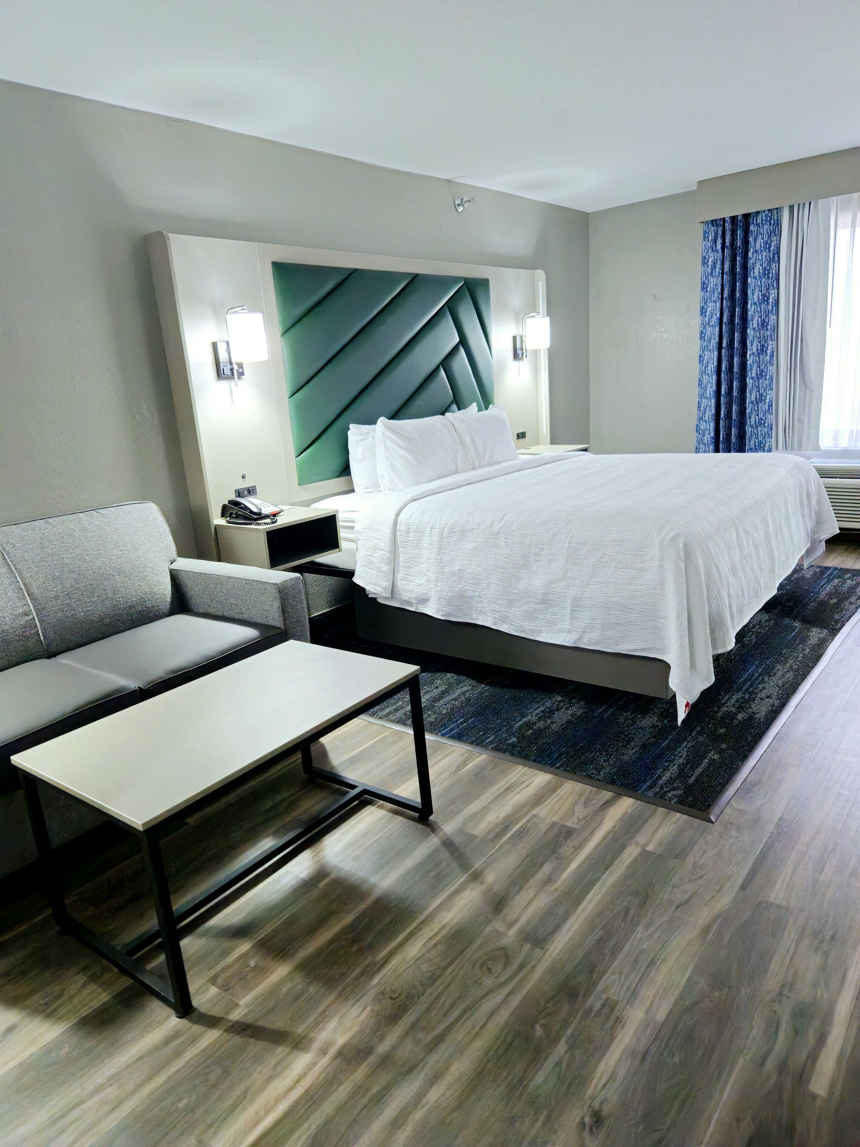 A modern hotel room with a bed, a sofa, a small table, and a wall-mounted light fixture.