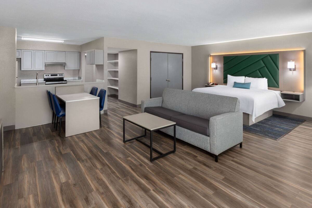 A modern hotel room with a kitchenette, a sofa, a bed, and a desk.