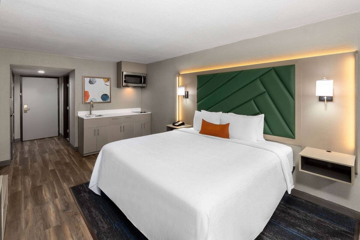 A hotel room with a large bed, a bedside table, a nightstand, a microwave, a sink, and cabinets.