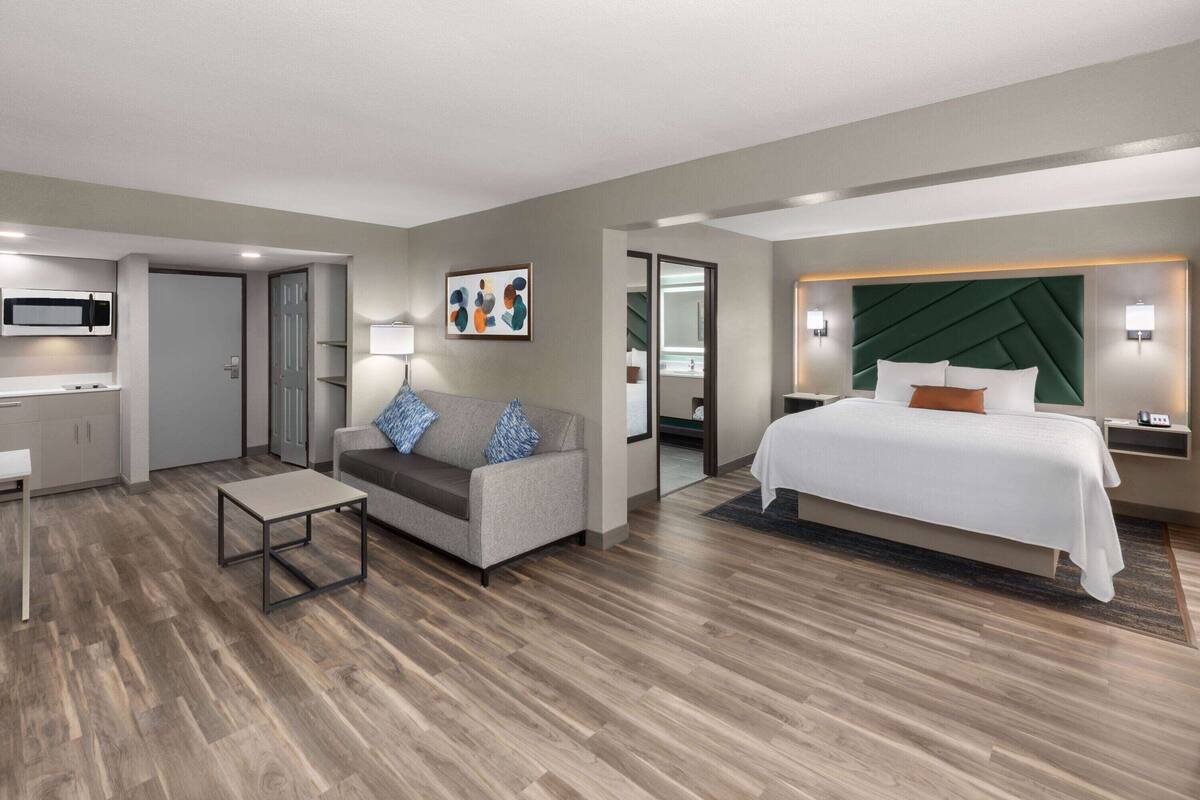 A modern hotel room with a bed, a sofa, a small table, a microwave, and a bathroom visible through an open door.