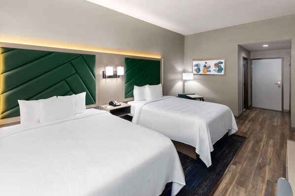 A hotel room with two beds, a green headboard, a nightstand with a phone, a lamp, and a painting on the wall.