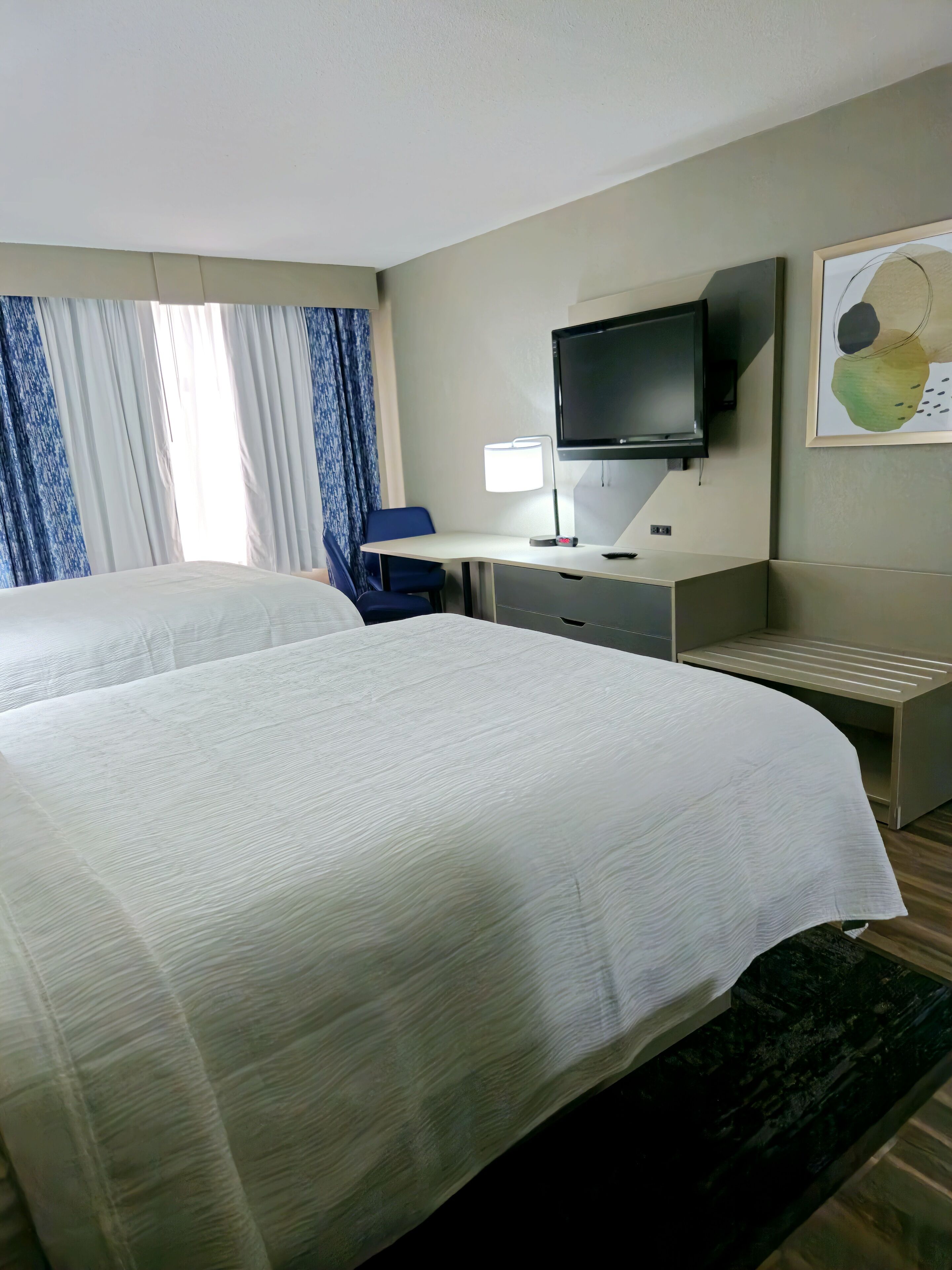 A hotel room with two beds, a desk with a lamp, a TV, and a painting on the wall.