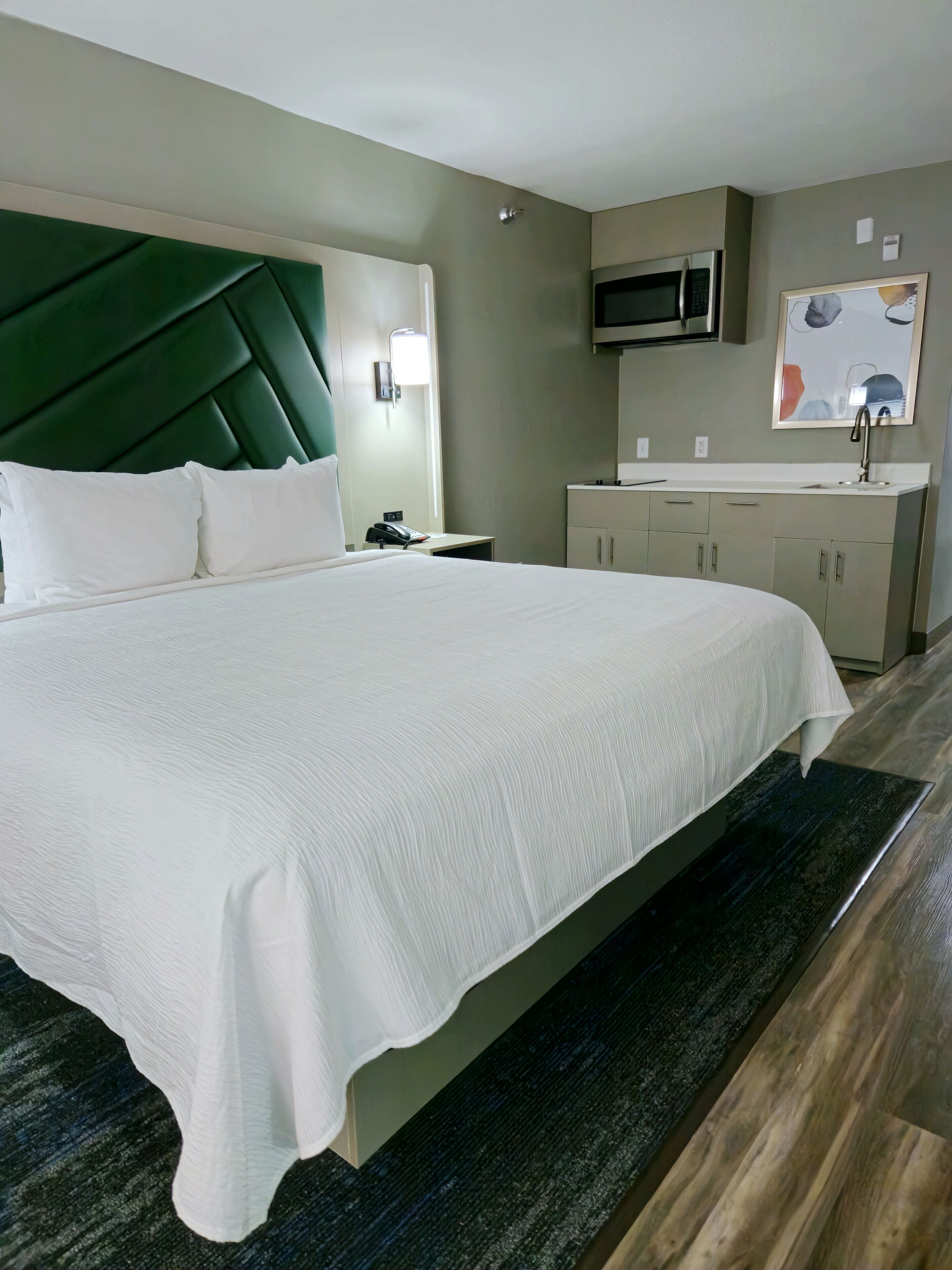 Ein Hotelzimmer mit einem großen Bett, einem Kopfteil, einem Nachttisch mit Lampe, einer Mikrowelle, einem Waschbecken und Schränken.