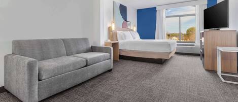 Una habitación de hotel moderna con un sofá gris, una cama con ropa blanca, un televisor de pantalla plana y un amplio ventanal con vistas al exterior.