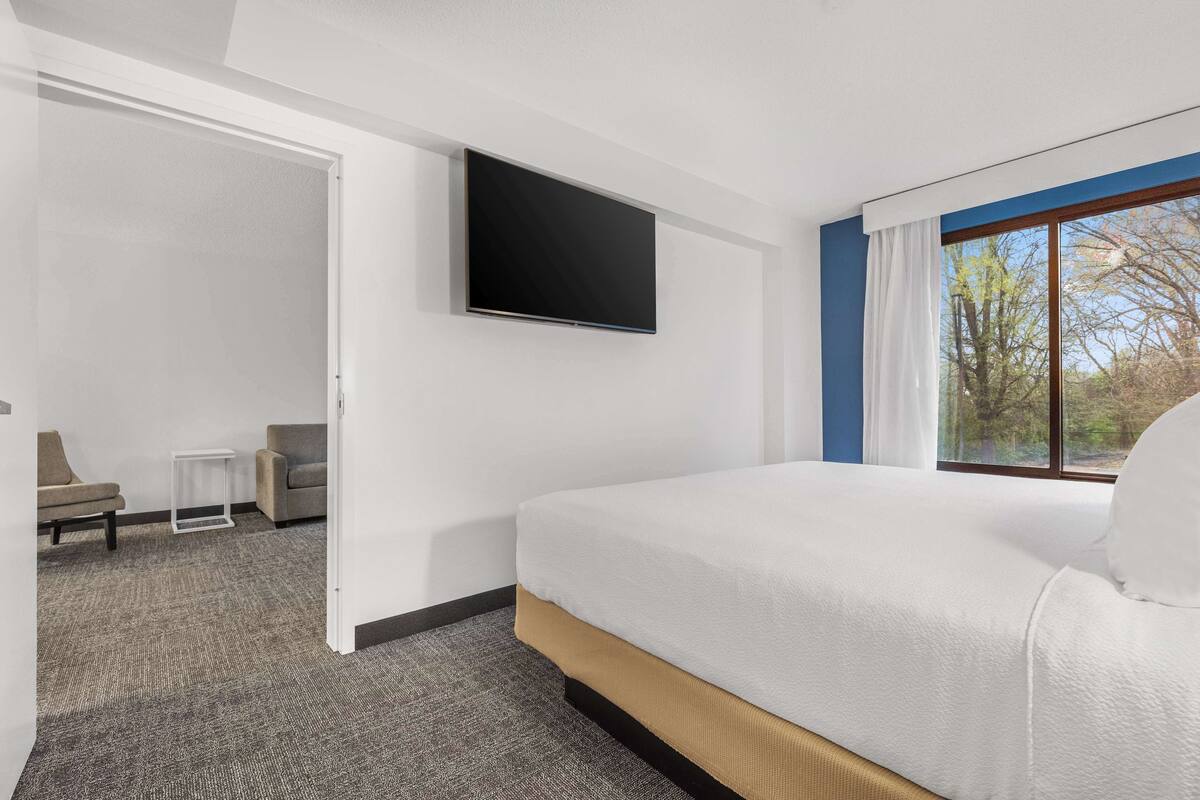 Ein modernes Hotelzimmer mit einem großen Bett, einem flachbildschirmfernseher an der Wand, einem kleinen Tisch und Blick ins Freie durch ein Fenster.