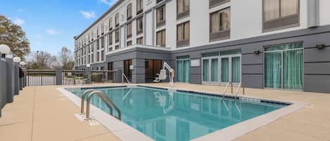 Un hotel moderno con piscina, mobiliario exterior y una amplia fachada.