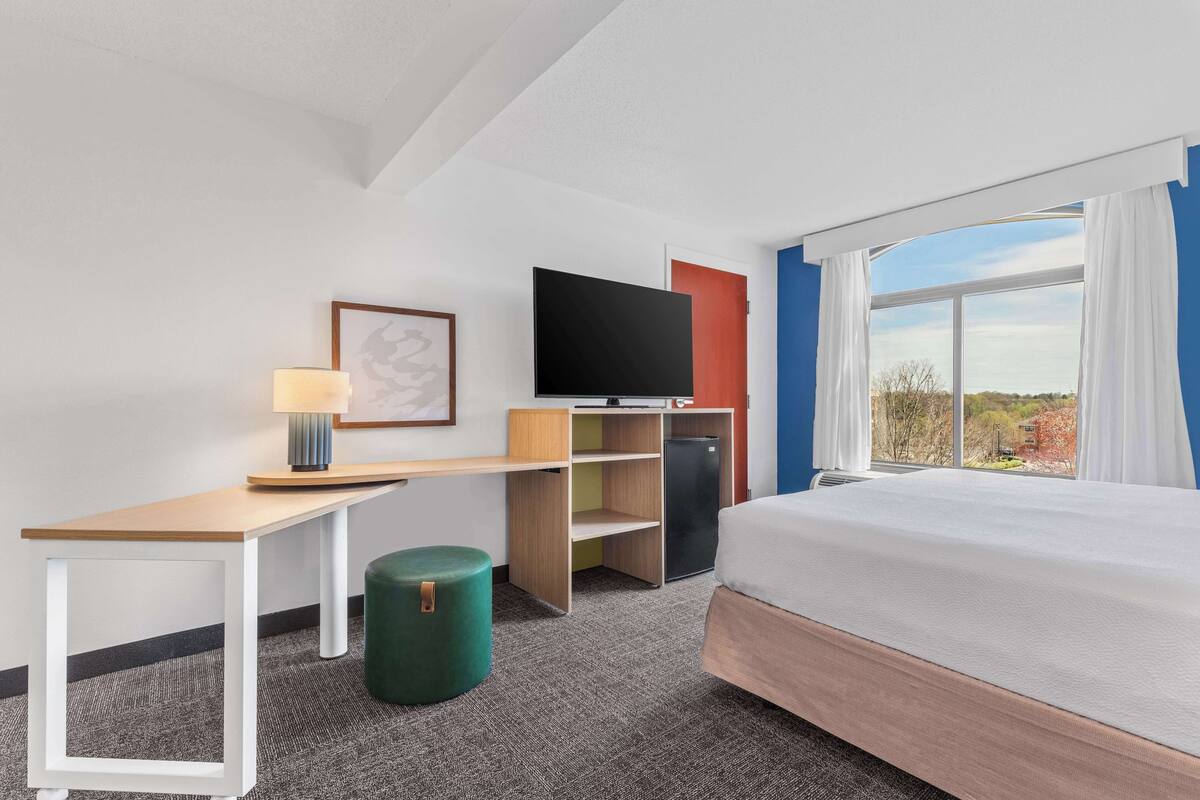 Ein Hotelzimmer mit einem Bett, einem Schreibtisch, einem Fernseher, einer kleinen Kühlschrank und einem Fenster mit Vorhängen.