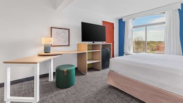 A hotel room with a bed, a desk, a television, a small refrigerator, and a window with curtains.