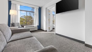 A modern hotel room with a grey sofa, a flat-screen TV mounted on the wall, and a large window with white curtains. - Spark by Hilton Charlotte Tyvola I-77 (Charlotte)