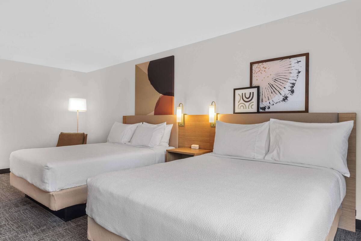 Ein Hotelzimmer mit zwei Betten, einem Nachttisch, einer Lampe und zwei gerahmten Kunstwerken an der Wand.