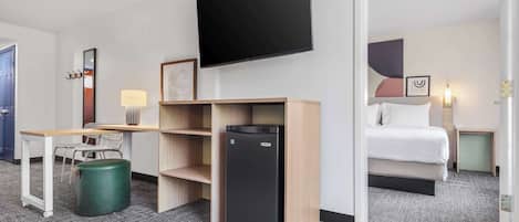 Habitación de hotel con cama, escritorio con silla, refrigerador pequeño, bote de basura, televisor montado en la pared y lámpara.