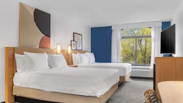 A hotel room with two beds, a desk, a TV, and a large window with a view of trees.