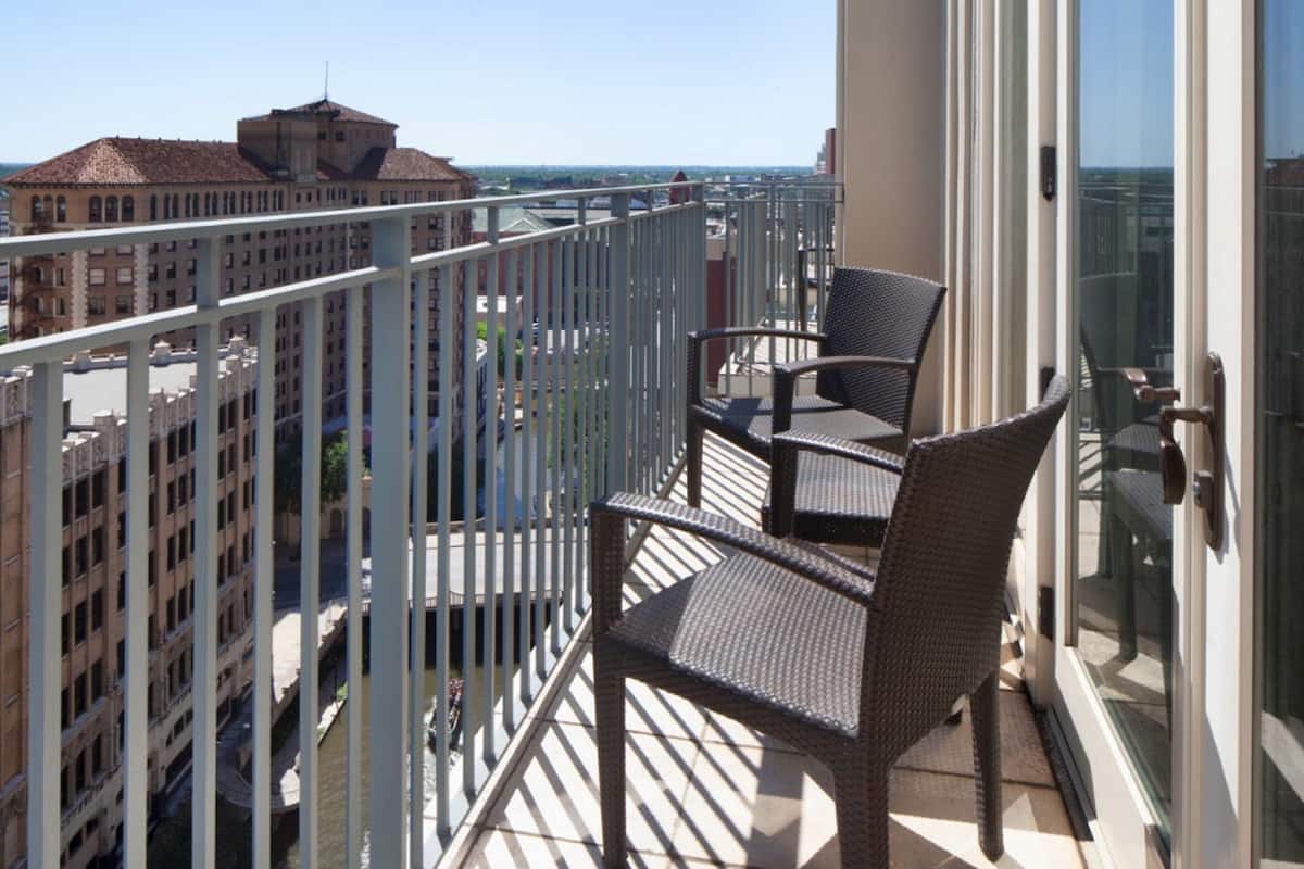 Een balkon met rieten stoelen en een glazen deur, met uitzicht op een stadslandschap met gebouwen en een rivier.