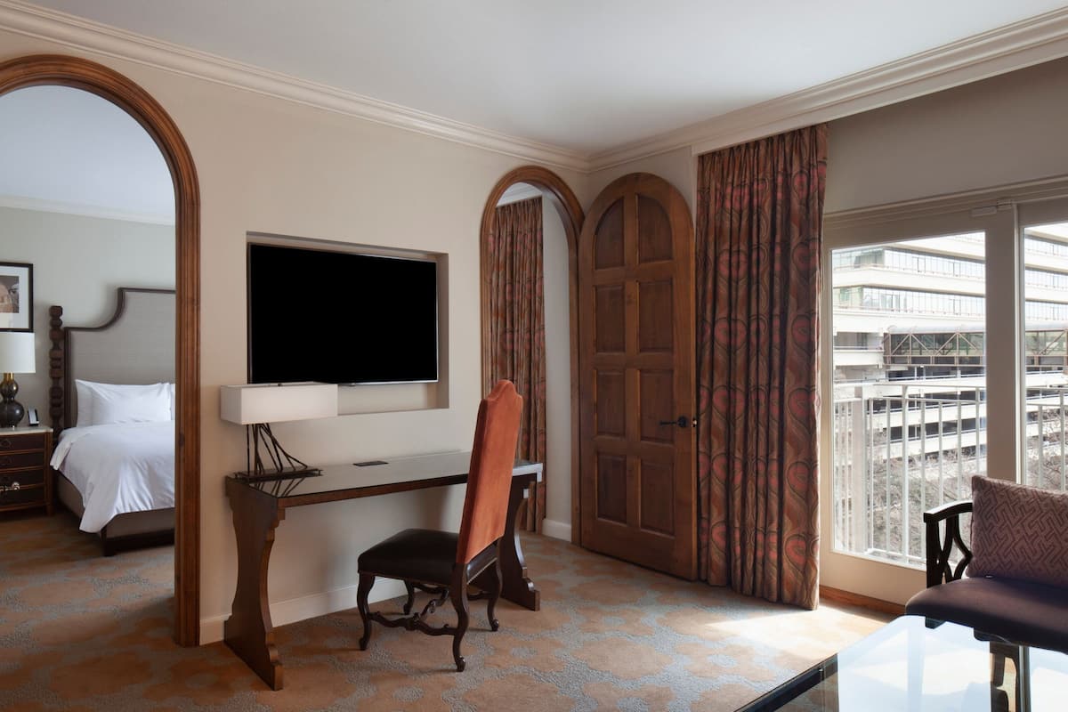 Een hotelkamer met een bed, een bureau met een televisie, een stoel en een groot raam met uitzicht op gebouwen.
