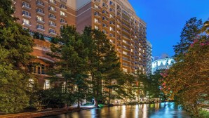 A multi-story hotel with balconies, a river in the foreground, and trees around.