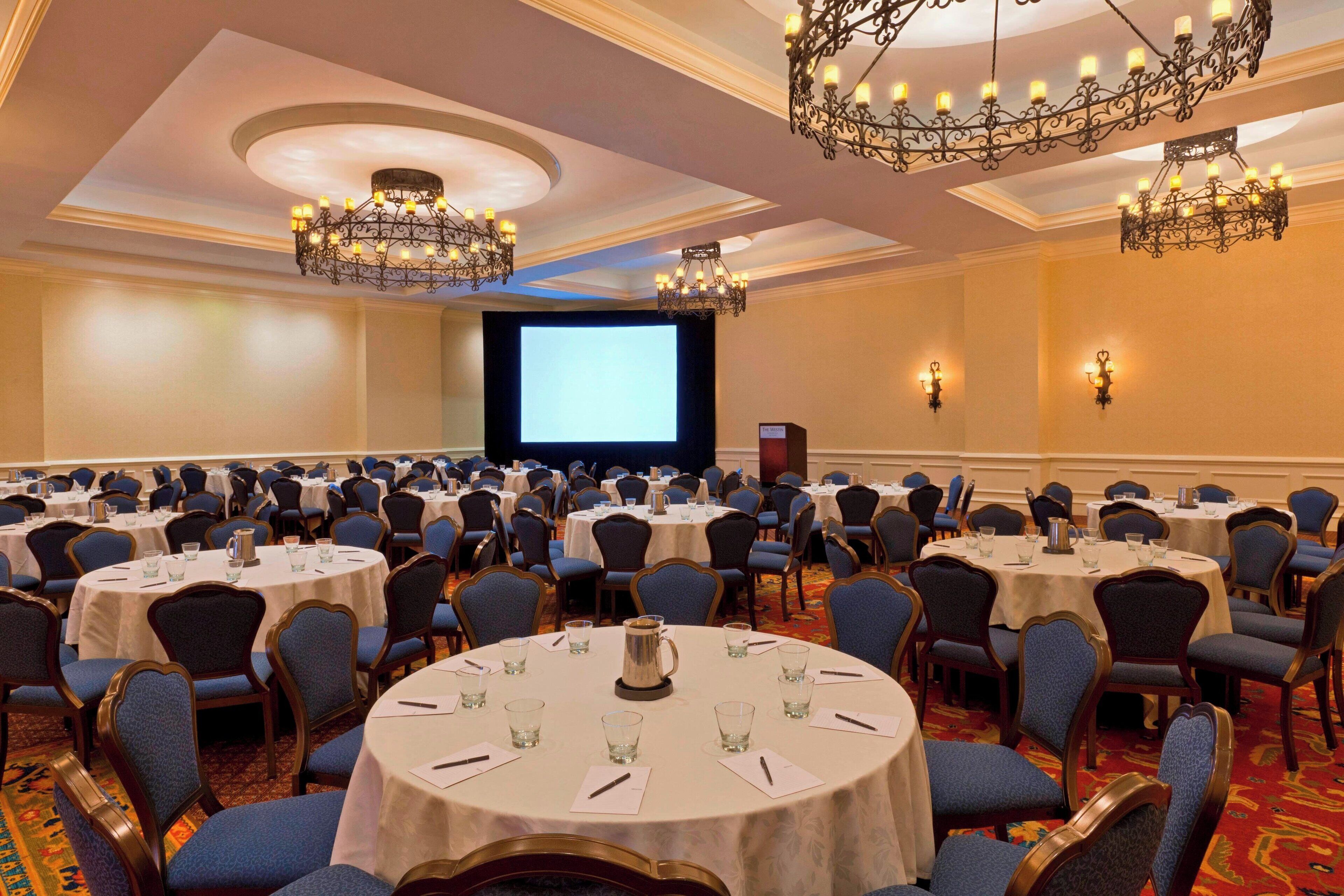 A conference room with round tables, blue chairs, chandeliers, and a projector screen.