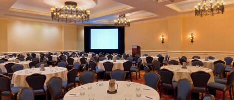 A conference room with round tables, blue chairs, chandeliers, and a projector screen.