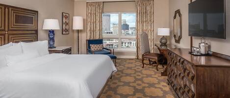 A hotel room with a large bed, a desk with a flat-screen TV, a chair, and a view of the city through the window.