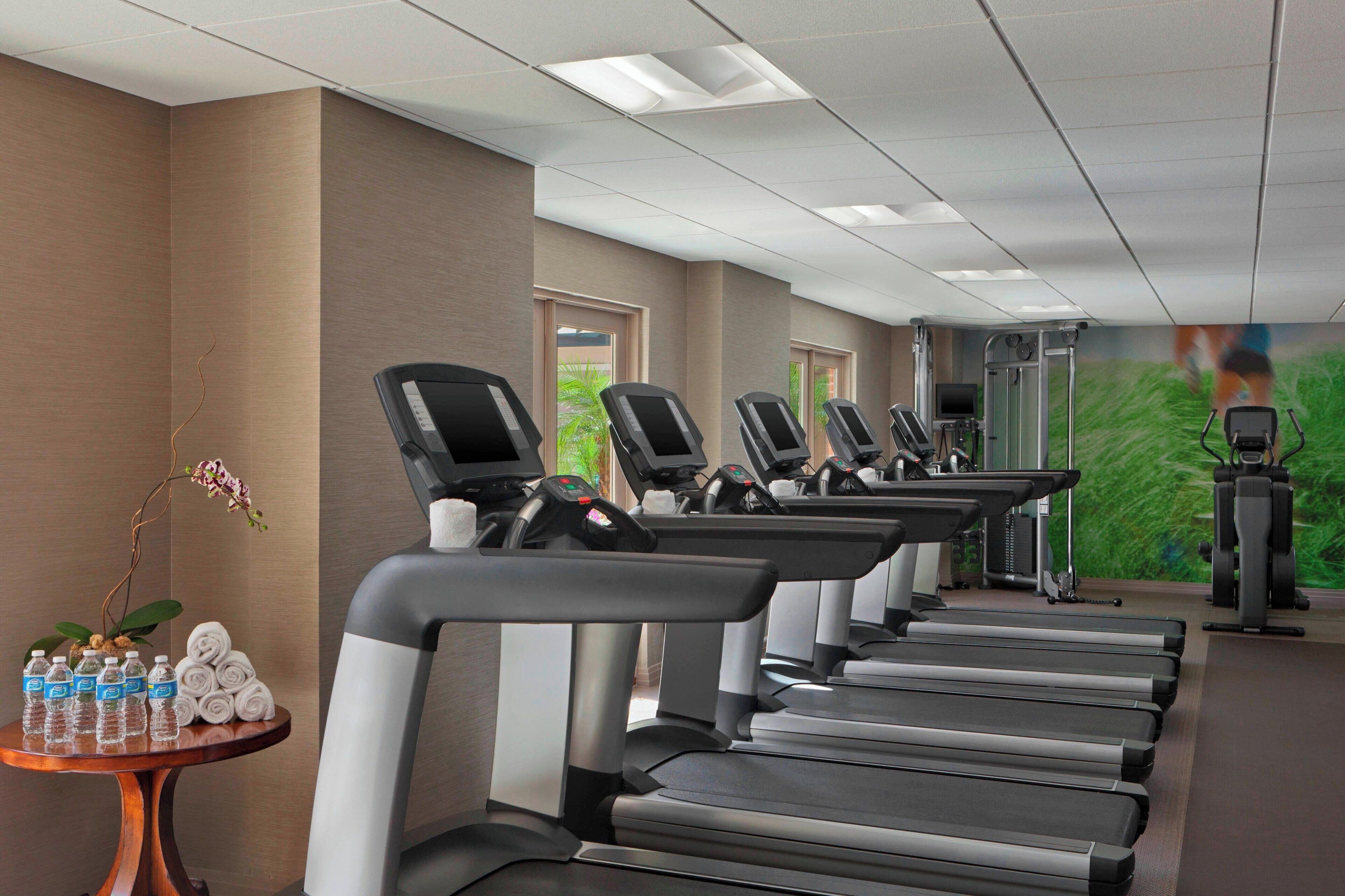 Varias cintas de correr en el gimnasio de un hotel, con un televisor de pared y una mesita con botellas de agua y toallas.
