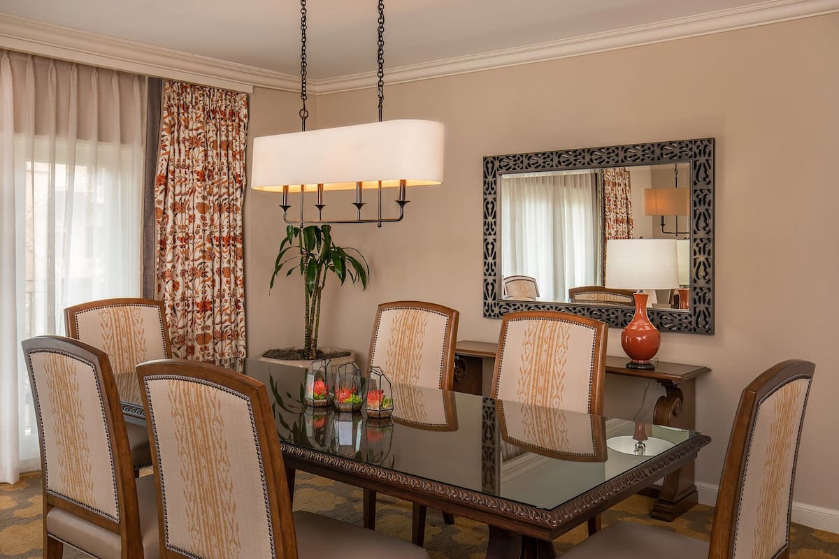 Een eetgedeelte met een glazen tafel, zes gestoffeerde stoelen, een hanglamp, een potplant en een decoratieve spiegel.