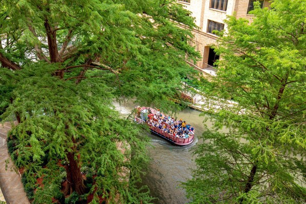Un paseo en barco por un río con pasajeros, rodeado de árboles verdes frondosos y un edificio al fondo.