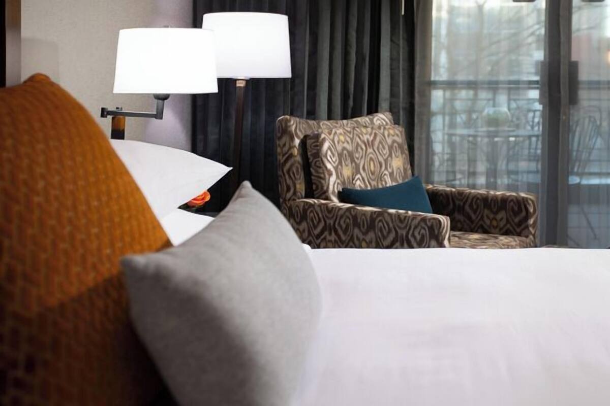 Quarto de hotel com uma cama, uma poltrona estampada, duas luminárias e vista pela janela.