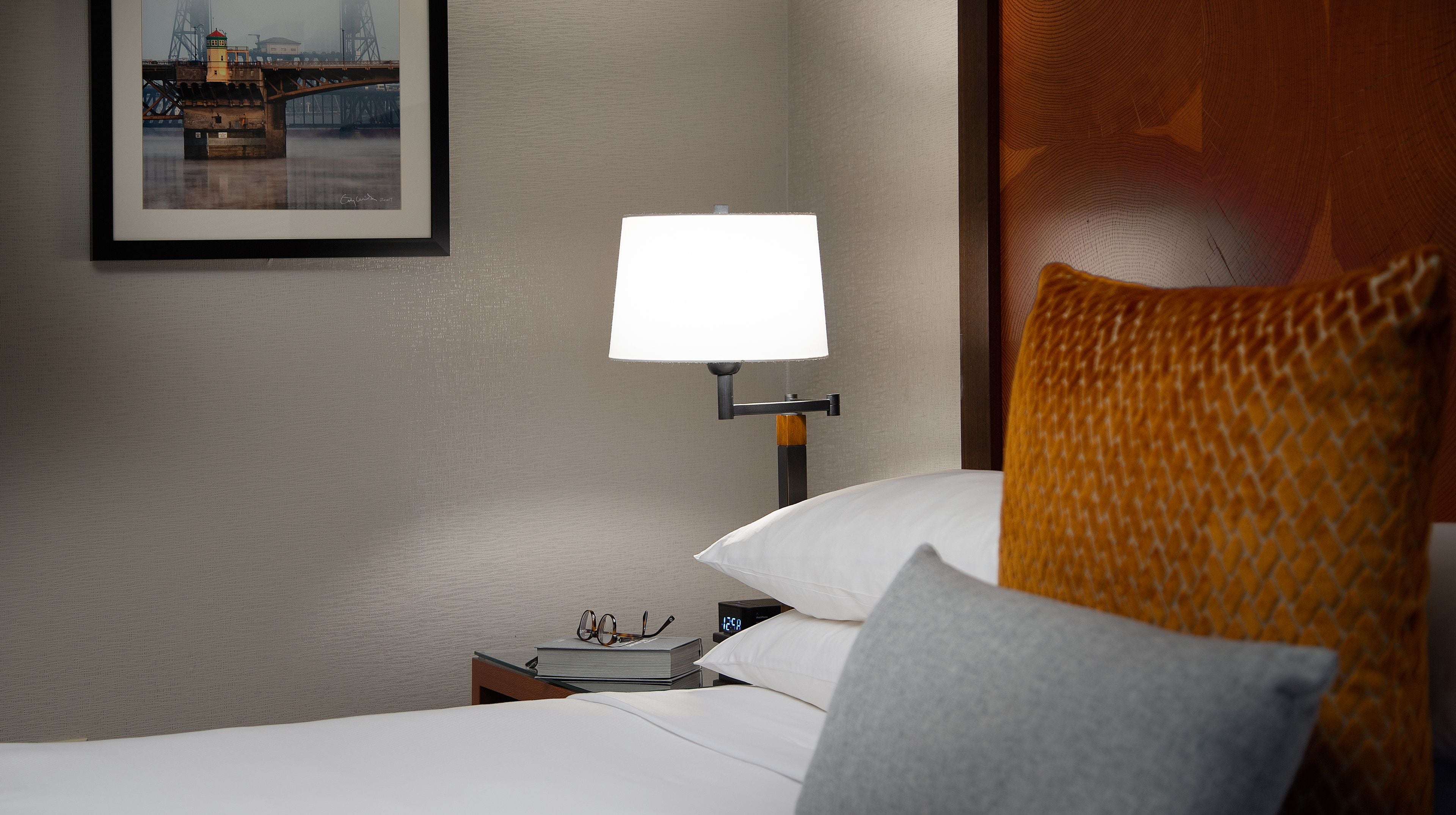 A hotel room with a bed, a nightstand with a lamp, a framed picture on the wall, and a digital clock.