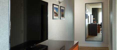 A hotel room with a flat-screen TV on a wooden dresser, a small table with a magazine, and a mirror reflecting a room with a desk and chair.