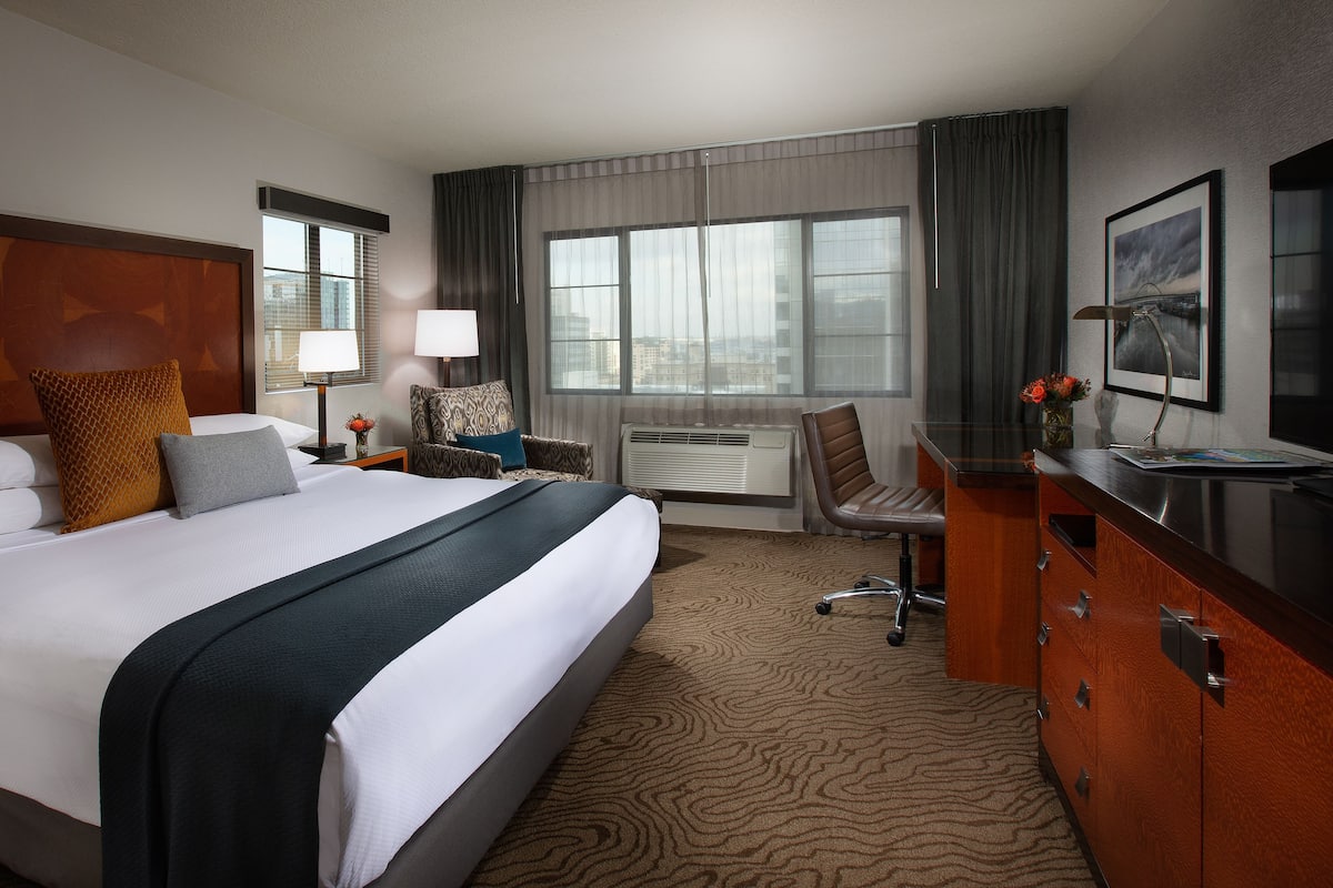 Quarto de hotel com cama grande, escrivaninha com cadeira, cômoda e janela com cortinas.
